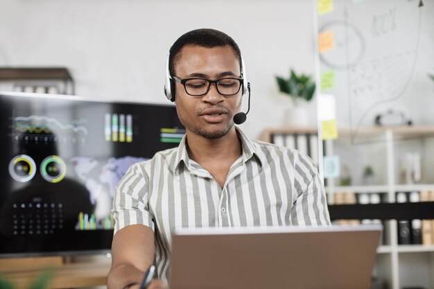 Confident african man in striped shirt using headset sitting at modern office and using laptop