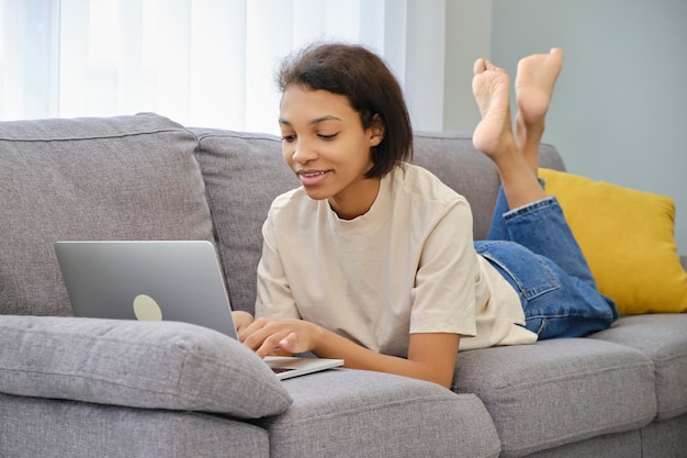 Confident africanamerican girl using laptop on smartphone sitting on sofa at home copy space