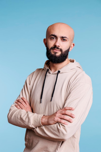 Confident arab man standing with crossed arms while looking at camera. Young serious arabian person wearing casual hoodie clothes posing with folded hands studio portrait