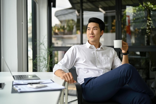 Photo confident asian businessman sits at his desk in modern office with a coffee cup