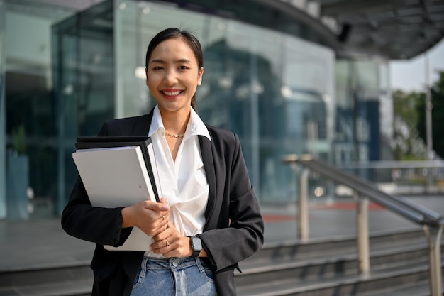 Уверенная азиатская деловая женщина, держащая папки с документами, улыбается, глядя в камеру