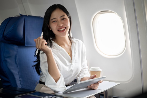 A confident Asian businesswoman is sitting at a window seat on a plane with her digital tablet