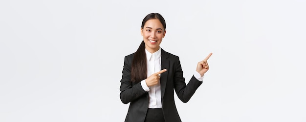 Confident asian female entrepreneur saleswoman in suit introduce product showing place to clients Businesswoman smiling and pointing fingers right at banner standing white background