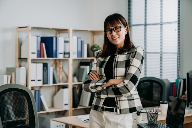 책상 위에 서 있는 자신감 있는 아시아 여성 기업가가 현대적인 밝은 개인 사무실에서 팔짱을 끼고 카메라를 바라보며 웃고 있습니다.