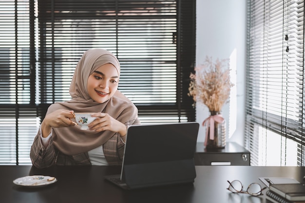 자신감이 아시아 이슬람 비즈니스 여자 갈색 hijab 앉아서 현대 사무실에서 랩톱 컴퓨터를 사용합니다.