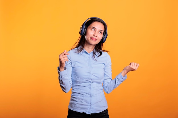 Confident asian woman listening music and dancing, enjoying break time in studio over yellow background. smiling model wearing headphones showing funny dance moves. entertainment concept