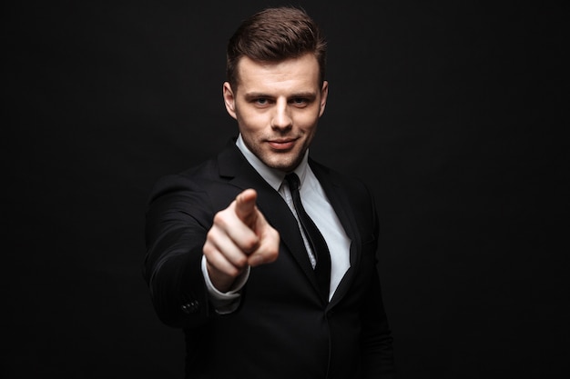 Confident attractive businessman wearing suit standing isolated over black wall, pointing finger at camera