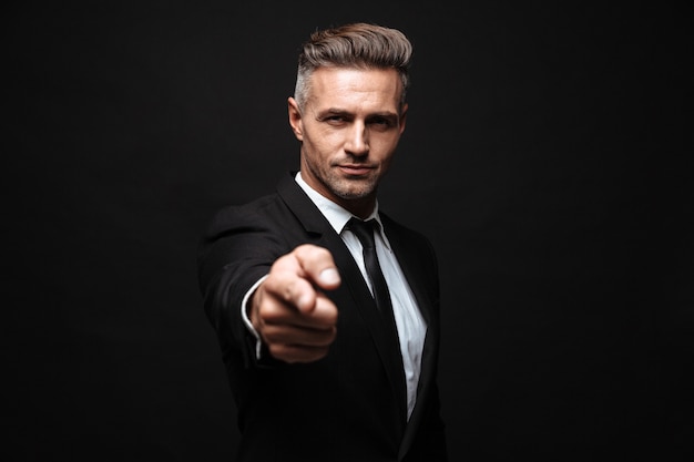 Confident attractive businessman wearing suit standing isolated over black wall, pointing finger at camera