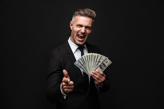 Confident attractive businessman wearing suit standing isolated over black wall, showing money banknotes, pointing finger