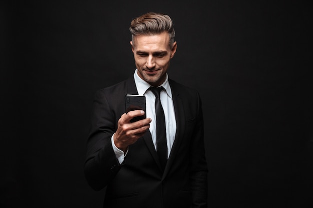 Confident attractive businessman wearing suit standing isolated over black wall, using mobile phone, celebrating success