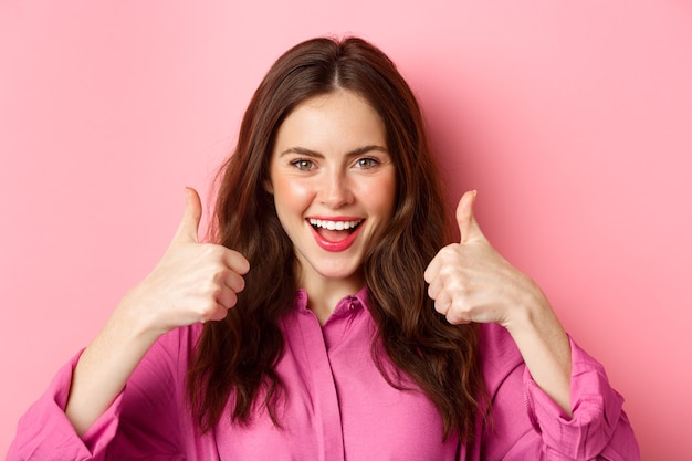 Confident attractive woman say yes, shows thumbs up with determined face, praise you, nice work gesture, standing over pink wall.