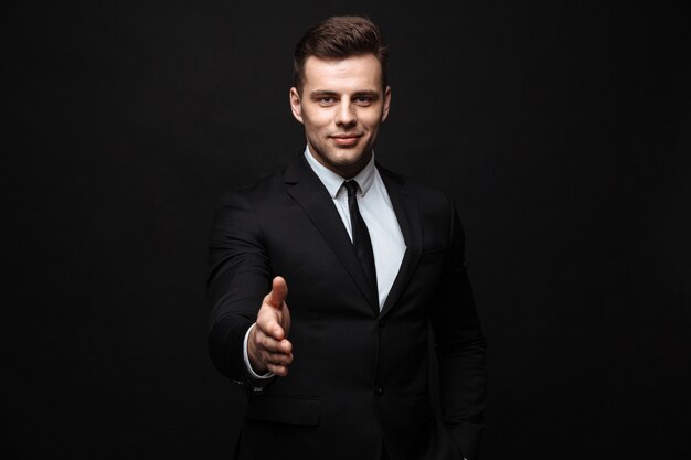 Confident attractive young businessman wearing suit standing isolated over black wall, holding outstretched hand for greeting