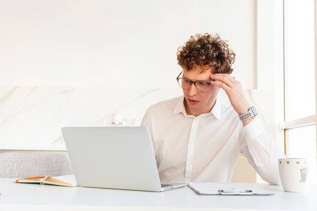 Confident attractive young businessman wearing white shirt working on laptop computer while sitting at the table at home, drinking coffee from a mug