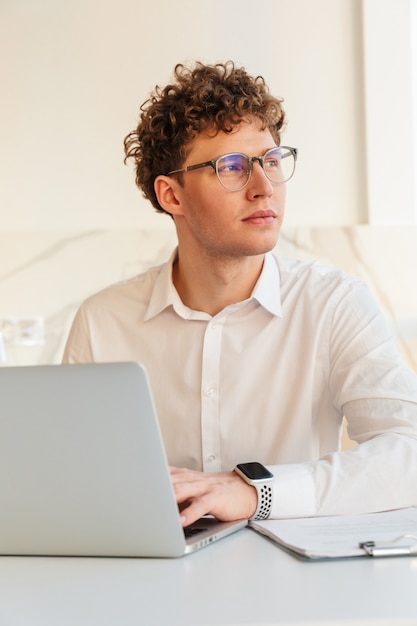 Confident attractive young businessman wearing white shirt working on laptop computer while sitting at the table at home