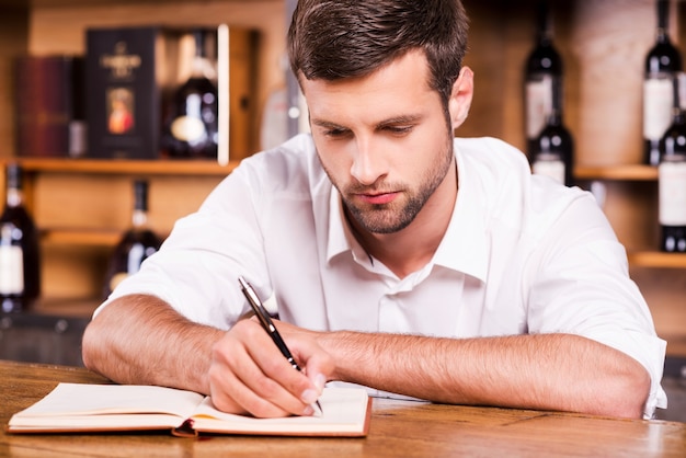 Foto proprietario del bar fiducioso. fiducioso giovane barista maschio in camicia bianca appoggiato al bancone del bar e scrivendo qualcosa nel blocco note