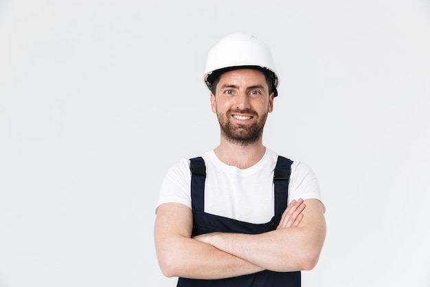 Confident bearded builder man wearing overalls and hardhat standing isolated over white wall, arms folded