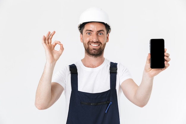 Confident bearded builder man wearing overalls and hardhat standing isolated over white wall, showing ok gesture and blank screen mobile phone