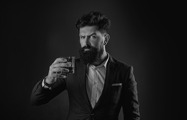 Уверенный в себе бородатый мужчина в черном костюме с бокалом виски на чердаке мужчина пьет алкоголь из стекла