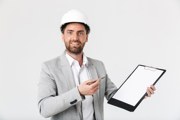 Confident bearded man builder wearing suit and hardhat standing isolated over white wall, showing blank notepad