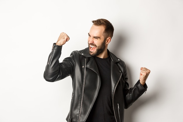 Confident bearded man celebrating victory, winning prize, making fist pump and rejoicing, wearing black leather jacket, white background.