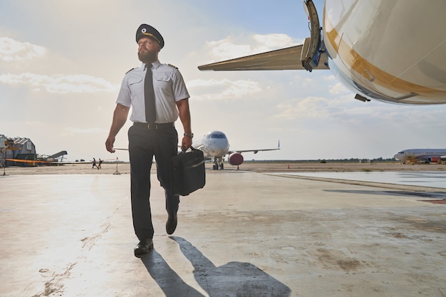 ブリーフケースを持って飛行機に向かって歩きながら遠くを見ている自信のあるひげを生やしたパイロット