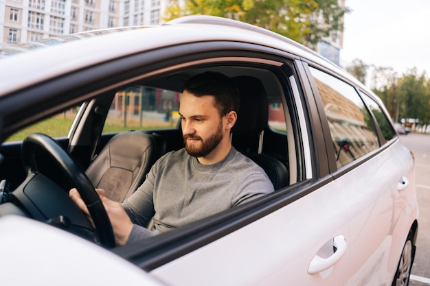 Confident bearded young man sitting in car and typing online message on cell phone side view