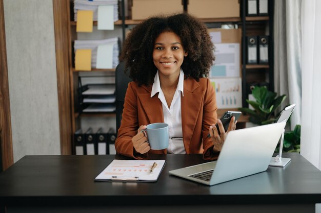 Photo confident beautiful african businesswoman typing laptop computer and digital tablet while holding coffee at modern office