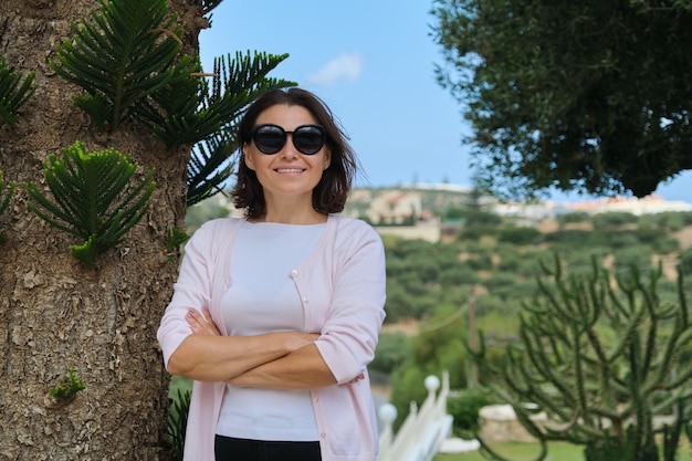 Confident beautiful mature businesswoman owner of tourist business, hotel posing with folded arms. Background resort landscape, tropical plants, copy space