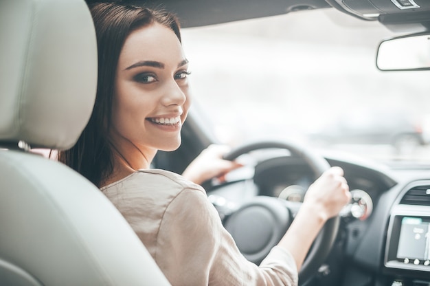 自信を持って美しい。車を運転しながら彼女の肩越しに見ているカジュアルな服装で魅力的な若い女性の背面図