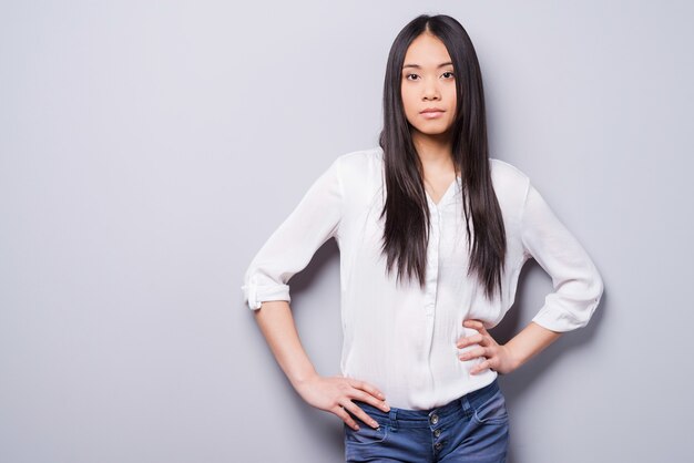 Confident beauty. Beautiful young Asian woman looking at camera and holding hands on hips while standing against grey background