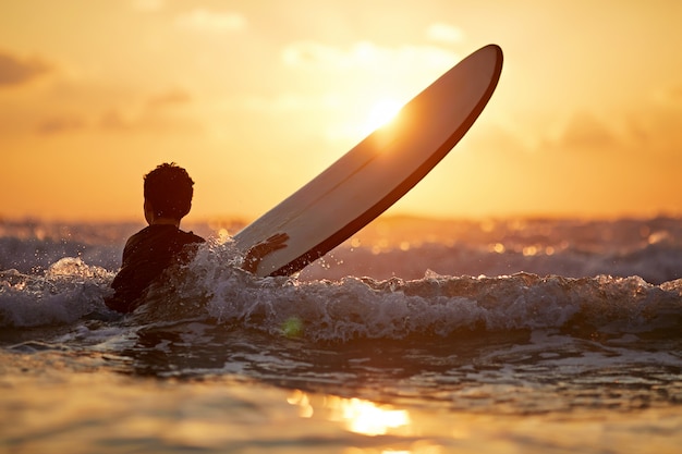 夕日の海辺に立っている間サーフボードを運ぶ自信の少年