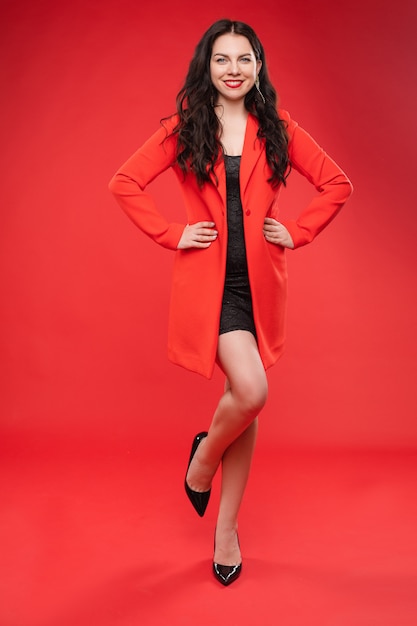 Confident brunette in black dress and red jacket posing