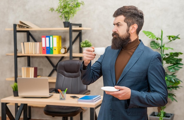Уверенный брутальный генеральный директор в деловом костюме обедает с чашкой кофе в офисе