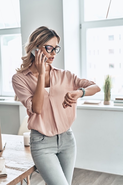 Confident business expert. Beautiful young woman talking on the mobile phone and checking the time while standing in modern office