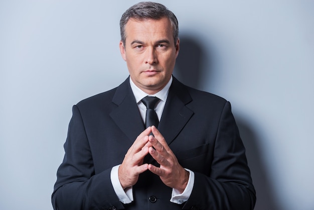 Photo confident business expert. confident mature man in formalwear holding hands clasped and looking at camera while standing against grey background