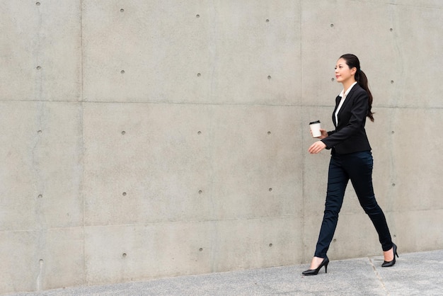 Photo confident business lady walking on gray wall background sidewalks outdoor corridor holding hot espresso coffee cup ready to go office meeting.