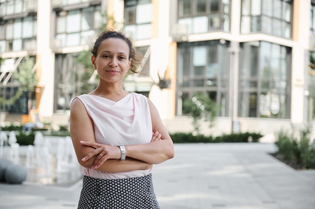 Confident business portrait of a successful middle aged business woman in casual attire posing to camera with crossed rams on the background of high buildings. Urban city backdrop