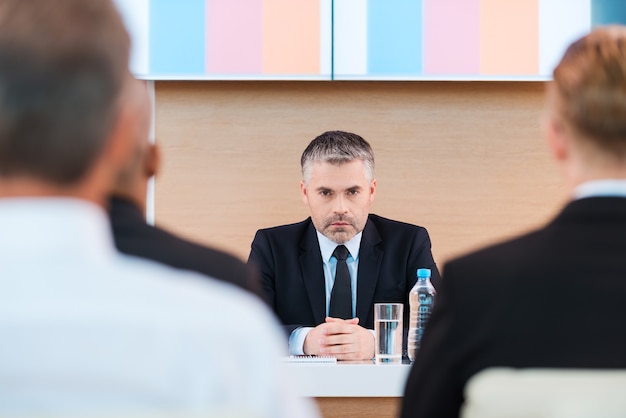 Confident business trainer. Confident mature man in formalwear sitting at the table with large monitor upon his head and with people on foreground