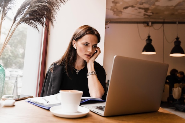 Confident business woman manager with laptop writes notes and working with colleagues online in cafe Attractive girl freelancer with cup of coffee is working online on laptop