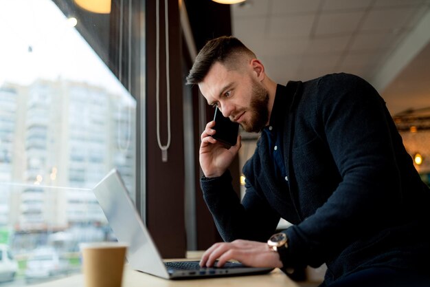 Confident businessman discusses issues on the phone and uses lap top in a cafe. Busy man working during coffee break. Closeup.