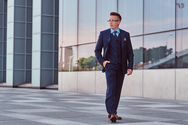 Confident businessman dressed in an elegant suit looking away and holds hand in pocket while walk on the street outdoors against cityscape background.