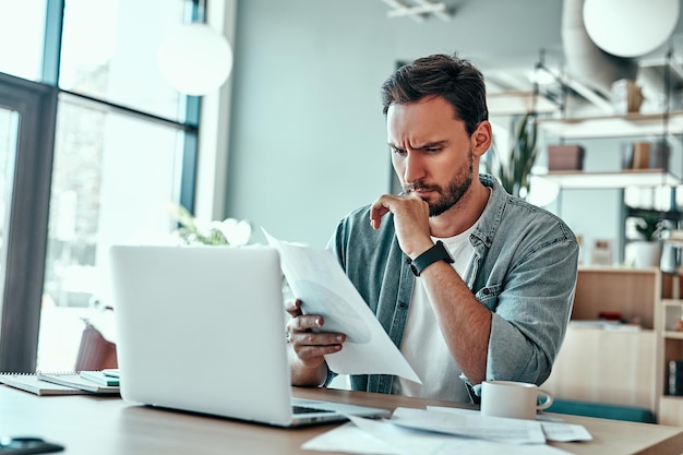 Confident businessman looking at financial documents in cafe Pensive man sitting in coffeeshop with laptop computer