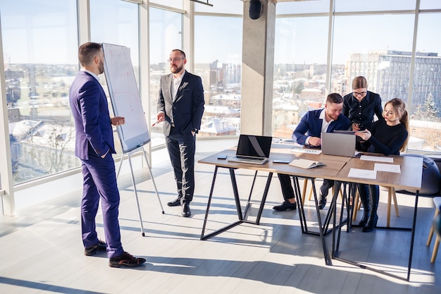 Confident businessman makes a presentation of a new project in the boardroom at a company meeting. Beautiful auditors talk with different partners about the business using a whiteboard and graphs.