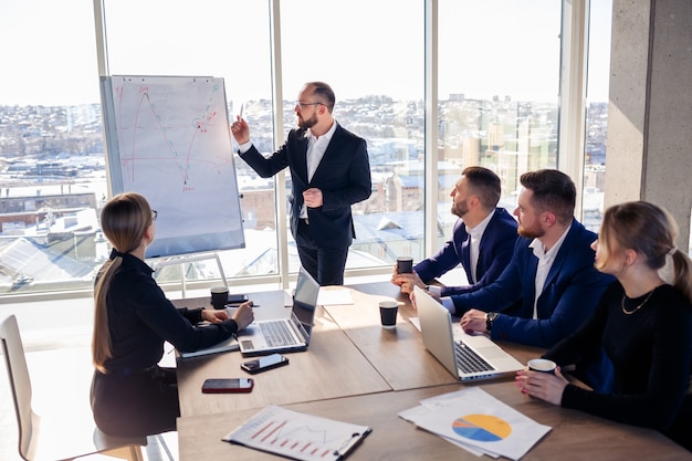 Confident businessman makes a presentation of a new project in the boardroom at a company meeting. Beautiful auditors talk with different partners about the business using a whiteboard and graphs.