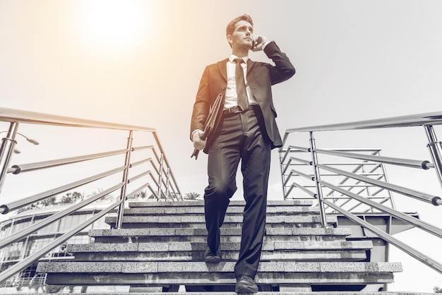 Photo confident businessman on the phone. low angle view of confident young man in formalwear talking on the mobile phone while moving down by staircase