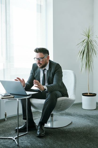 Confident businessman sitting in front of laptop in office and communicating with business partners through video chat