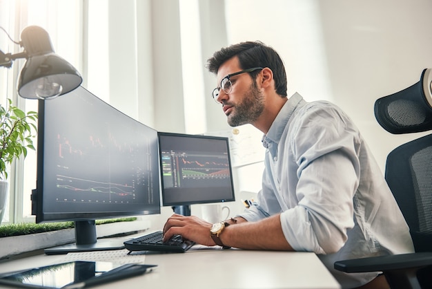 Photo confident businessman. young bearded trader in formal wear is analyzing trading charts on computer screens while sitting in his modern office