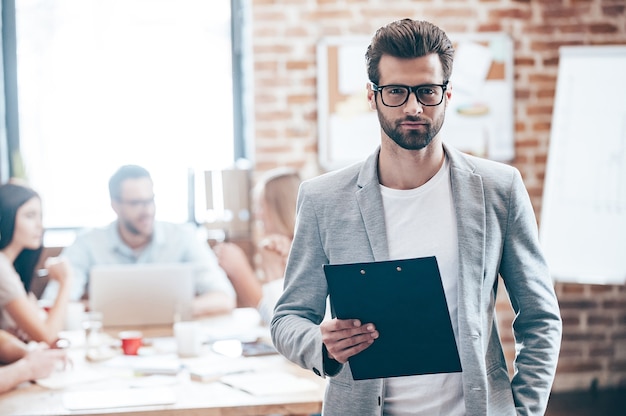 Confident businessman. Young handsome man holding notepad and looking at camera while his colleagues discussing something in the background