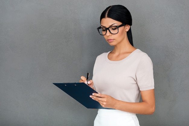 Confident businesswoman. Confident young businesswoman writing in clipboard while standing against grey background