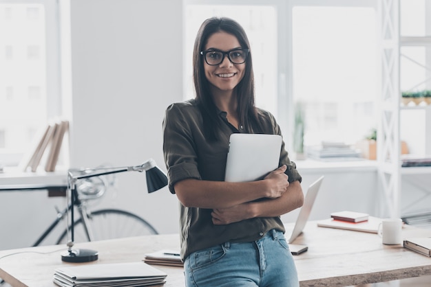 Confident businesswoman. Confident young woman in smart casual wear holding digital tablet and smiling while standing near her working place